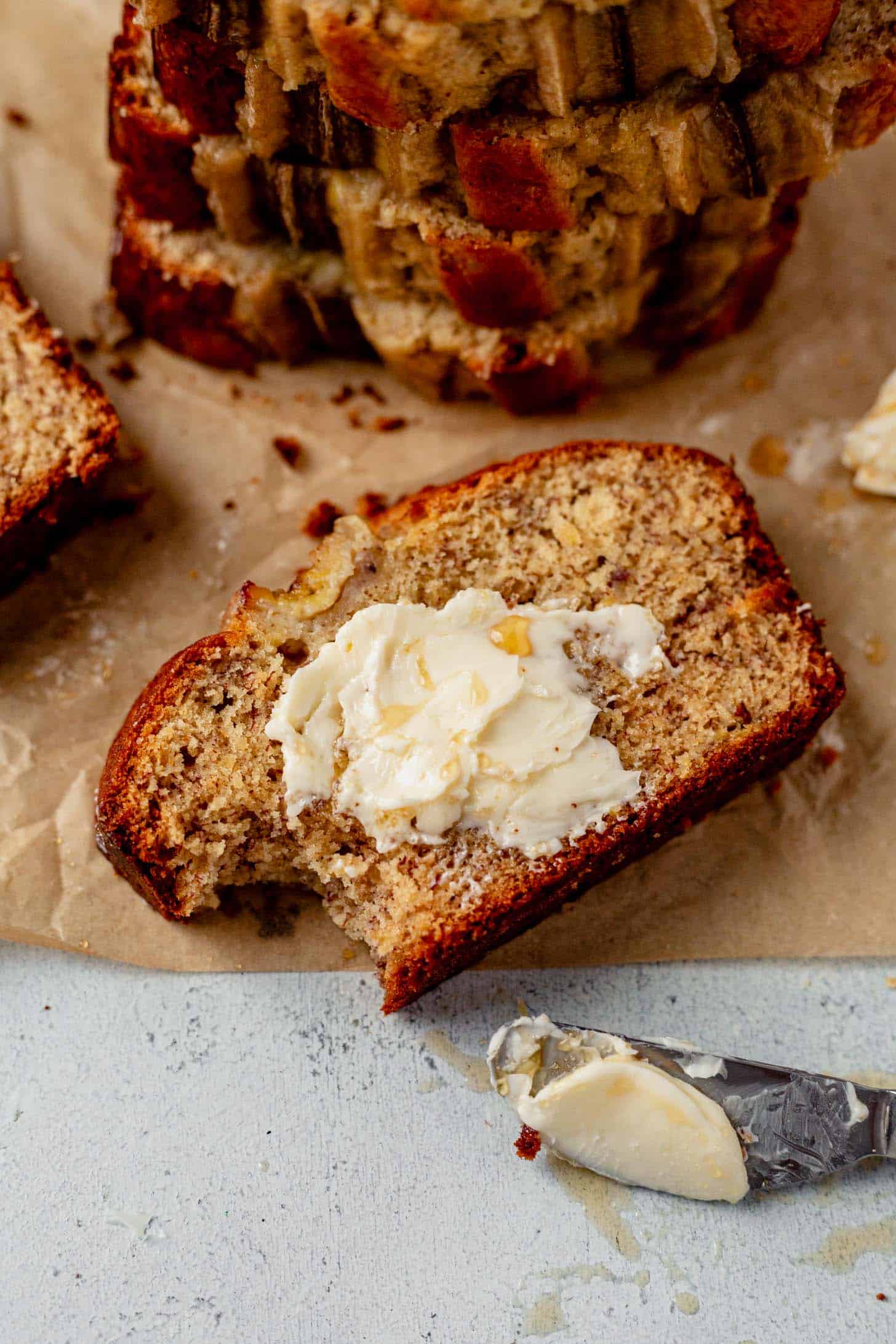 Close-up of a slice of banana bread with a bite taken out, topped with softened butter and drizzled honey, resting on parchment paper beside a butter-smeared knife.