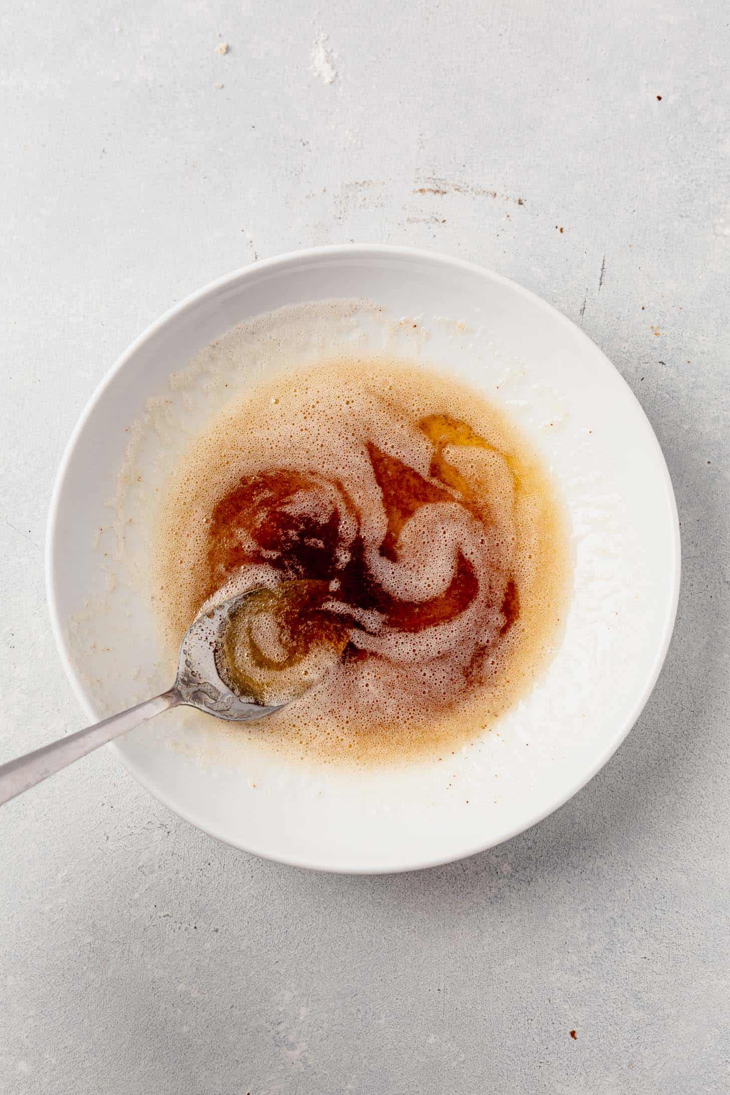 Swirled browned butter in a white bowl with a spoon, showing golden foam and deep amber hues on a light gray surface.