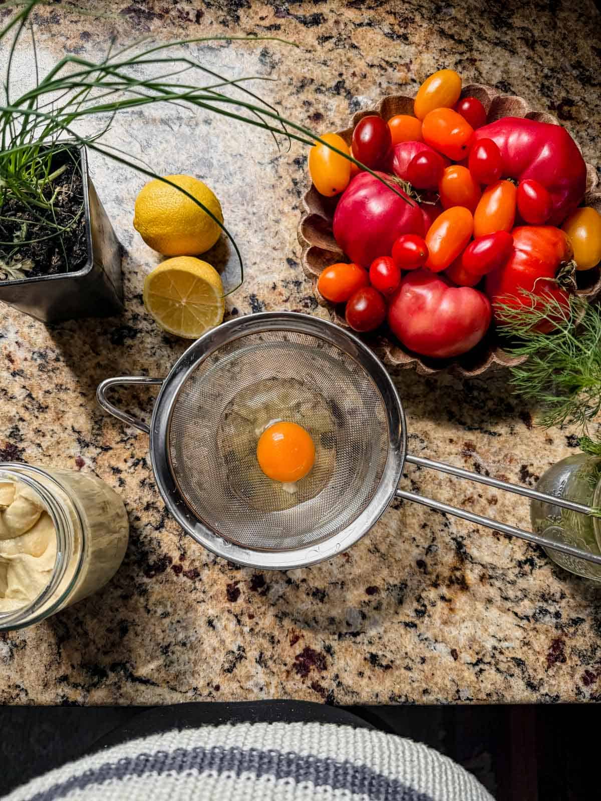 straining a raw egg through a mesh strainer next to a bowl of tomatoes, lemon, and chives.